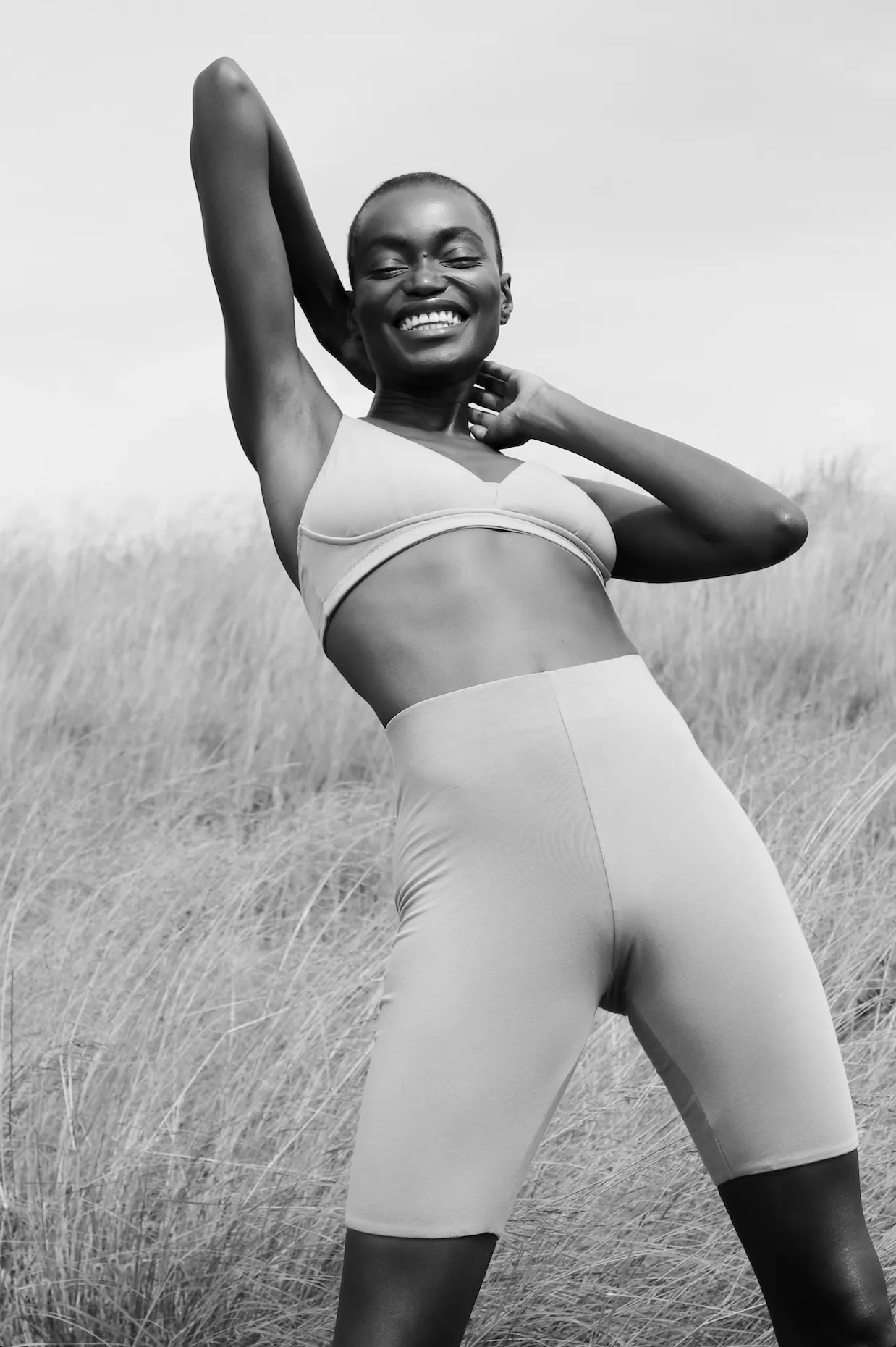 Black and white photograph of a model in a grey bralette and bike shorts, arm raised, in tall grass