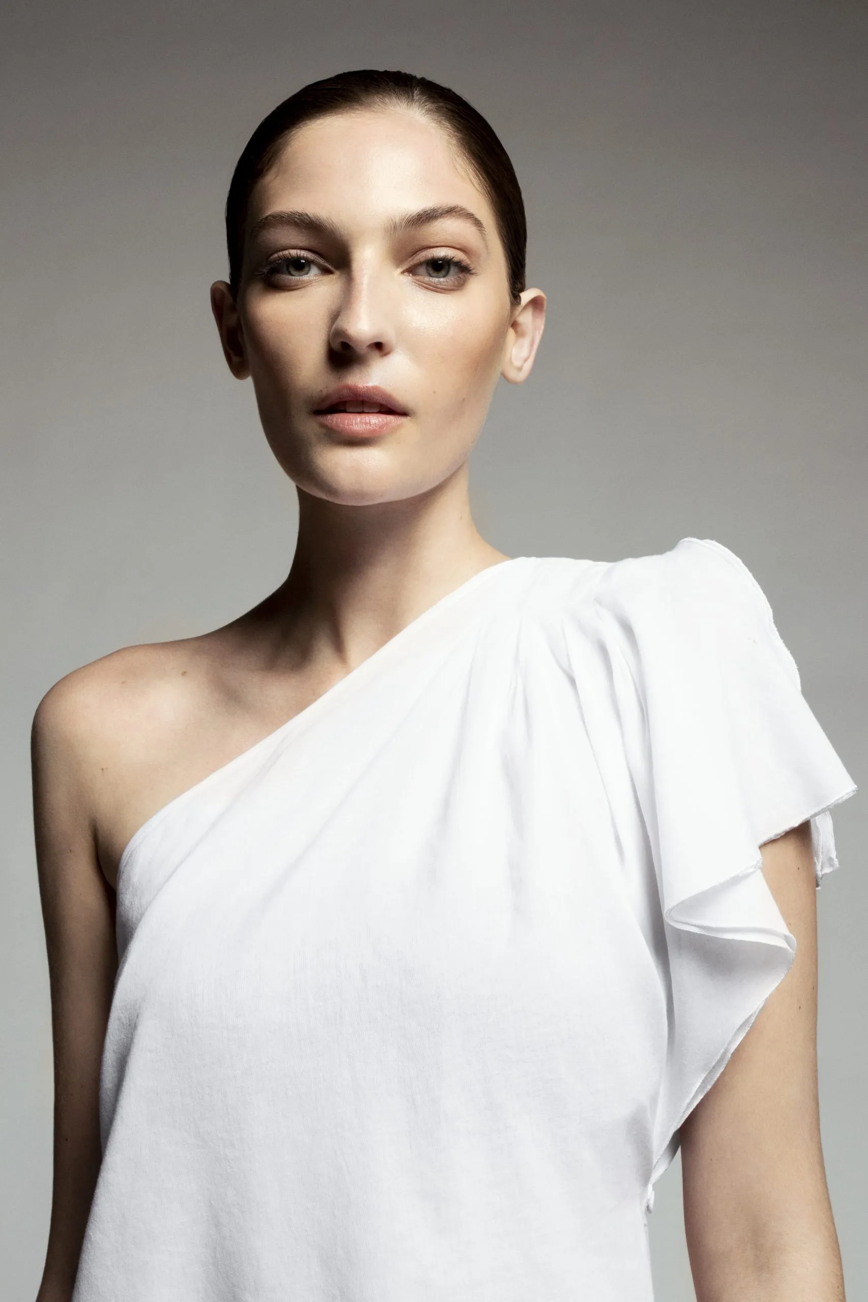 Model in a white one-shoulder top with a draped ruffle sleeve, photographed against a grey studio backdrop