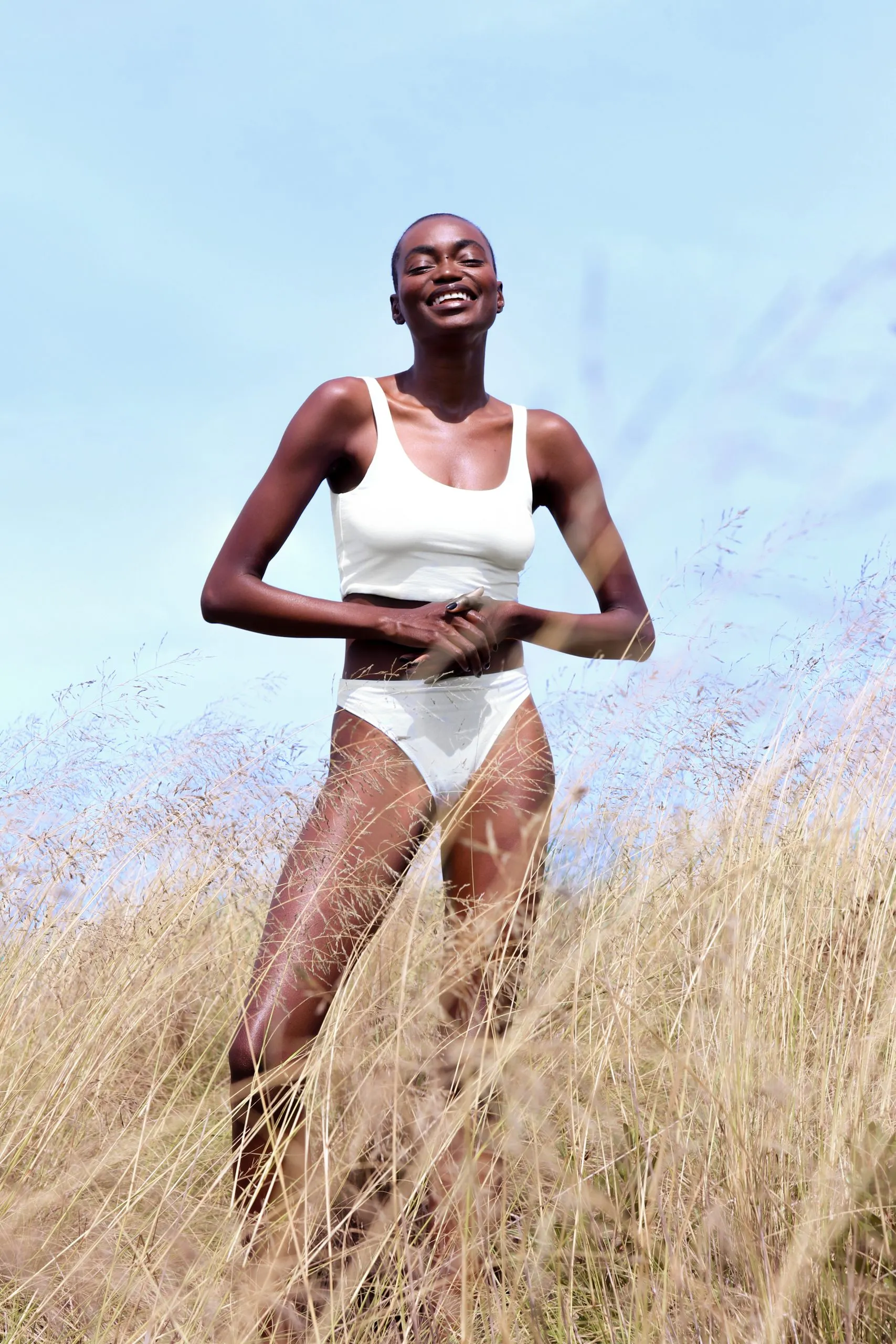 Model laughing in a white two-piece, standing in tall golden grass under a pale blue sky